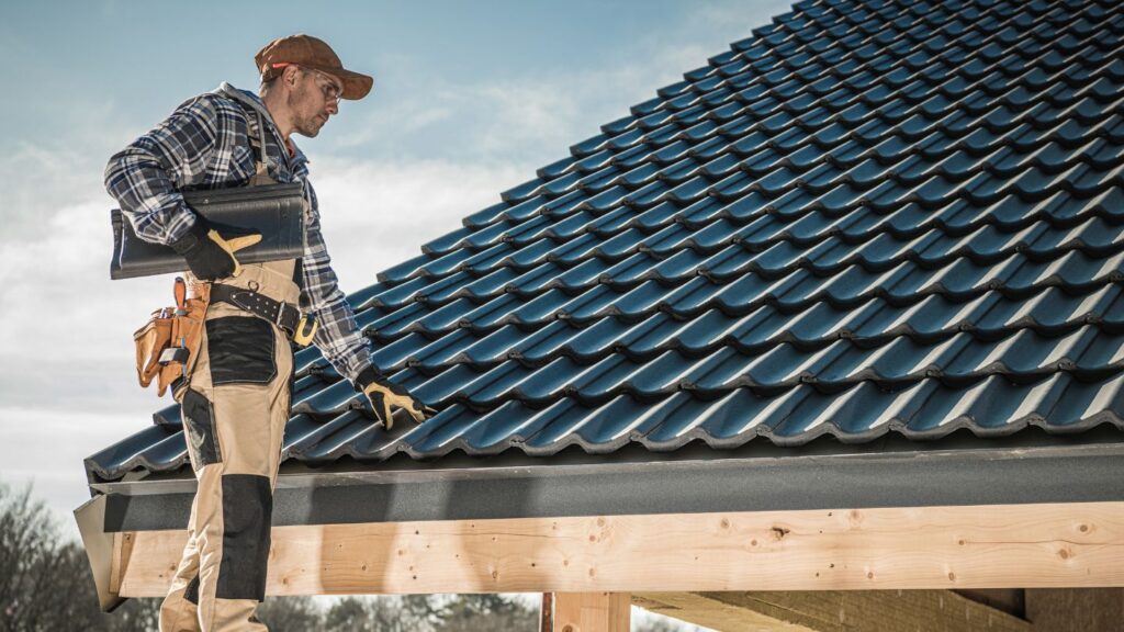 roofer wearing safety gears carrying metal tiles with a house with a metal roof in the background