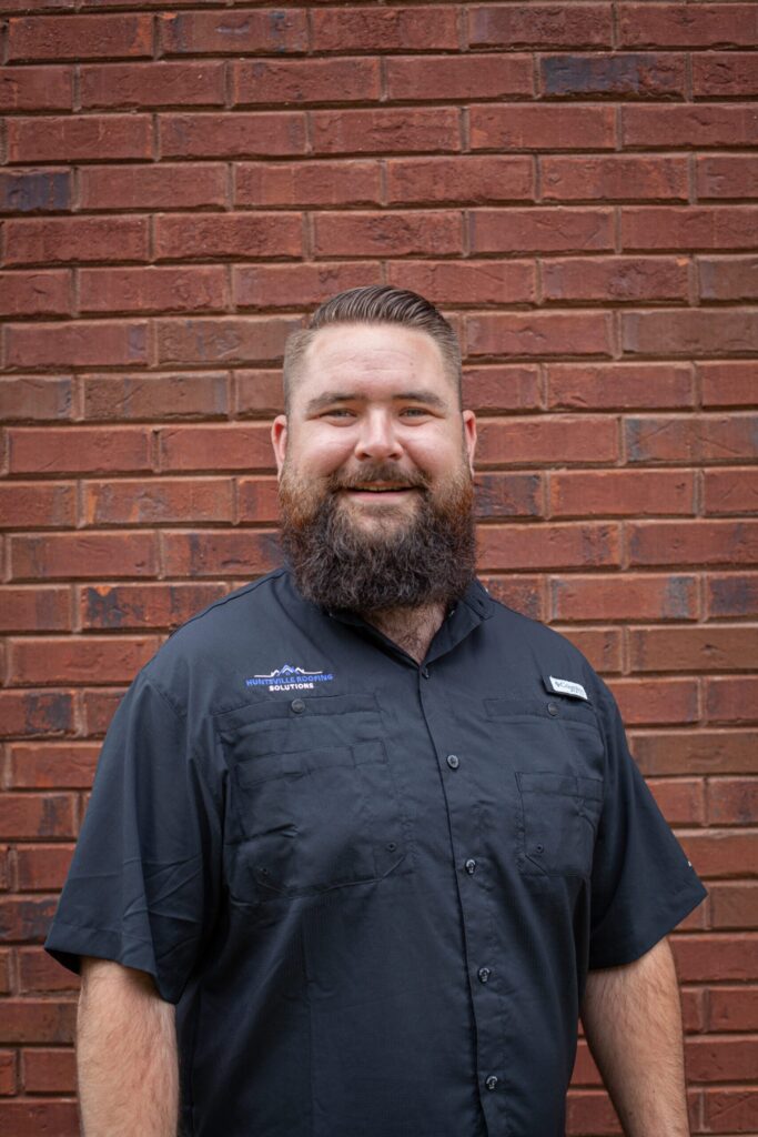 Barry Minor of Huntsville Roofing in a black t-shirt, smiling, with brick exterior walls in the background