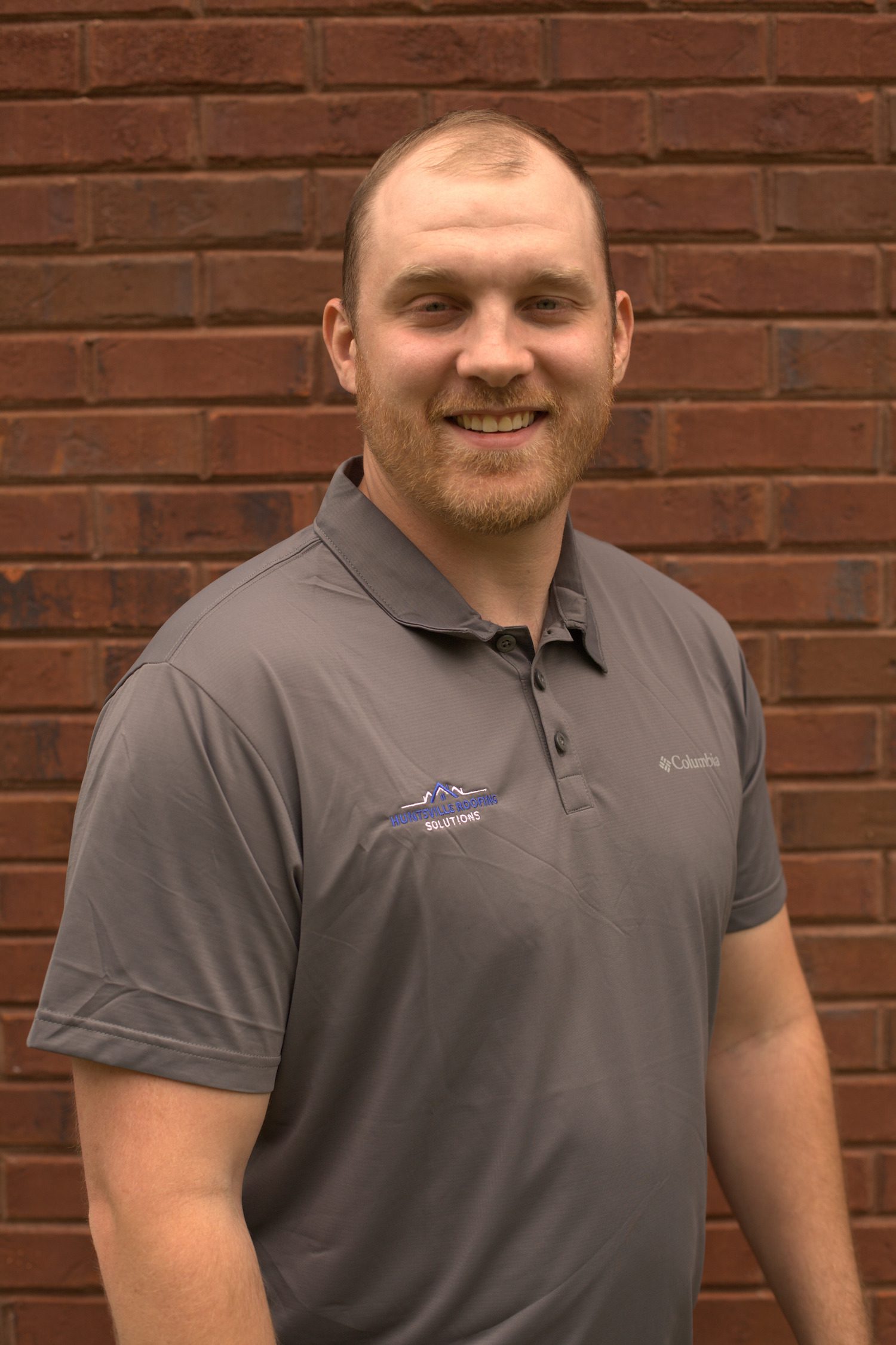 Jordan Woolf of Huntsville Roofing in a grey t-shirt, smiling, with brick exterior walls in the background