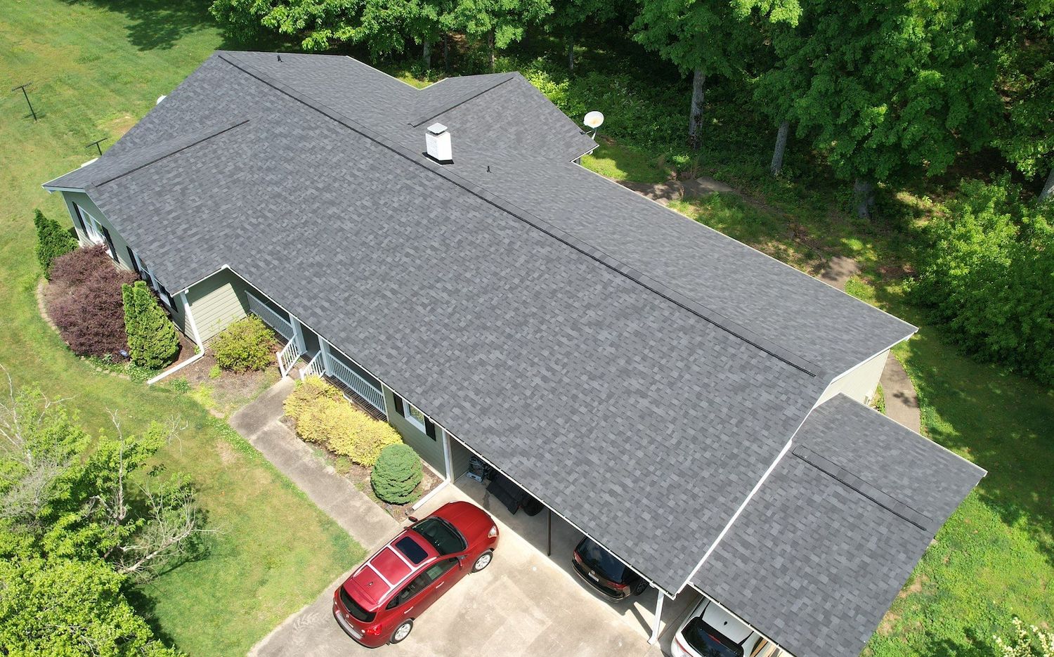 drone view of a house with granite grey architectural asphalt shingle roof and red car in the driveway with green lawn and trees in the surrounding